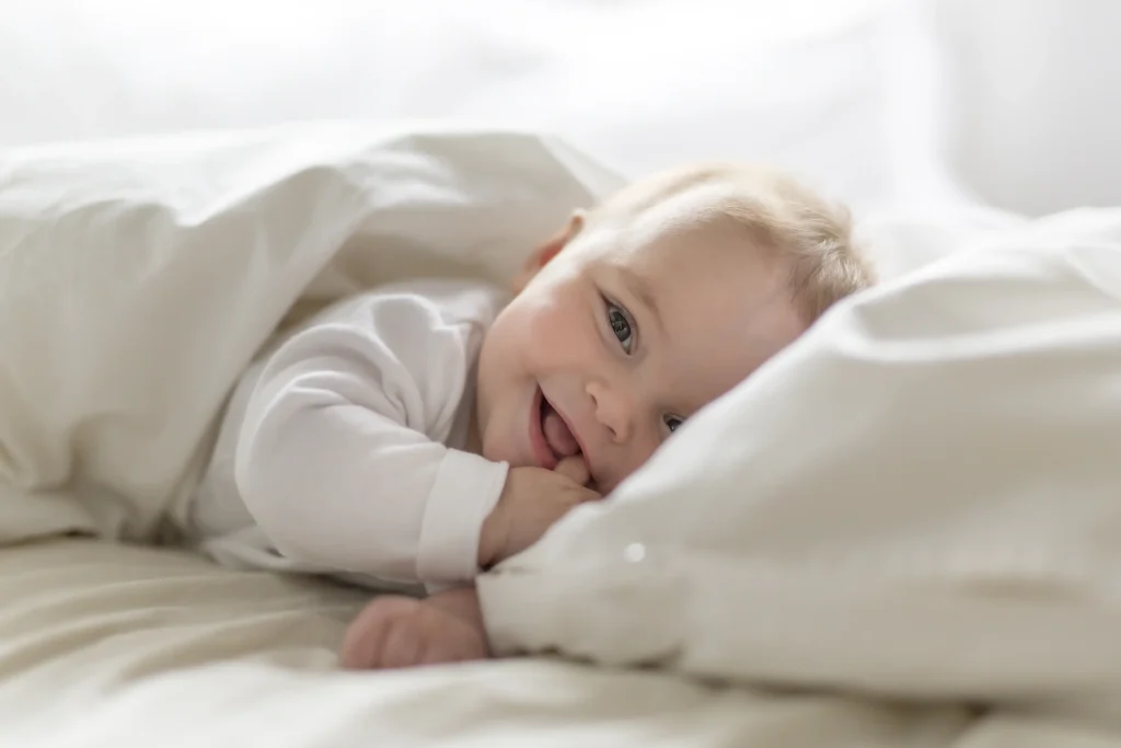 A baby smiles looking at the camera with their thumb in their mouth while lying in soft white bedding.