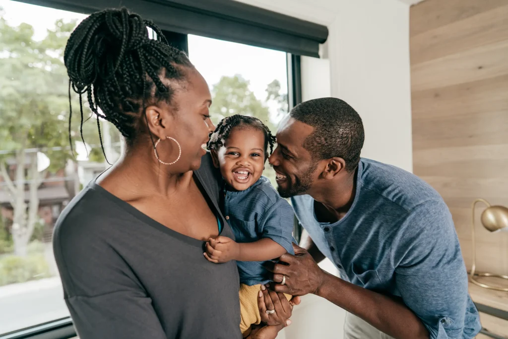 A couple stands near a window, the woman holds a laughing toddler while the smiling man leans in