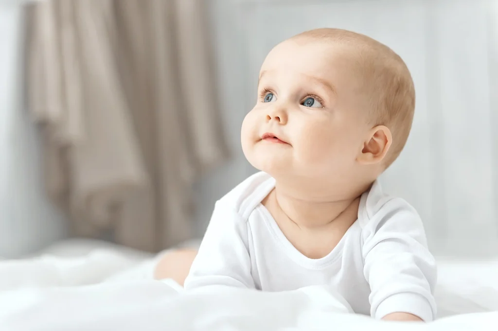 A baby crawls on soft white bedding while smiling and looking upwards.