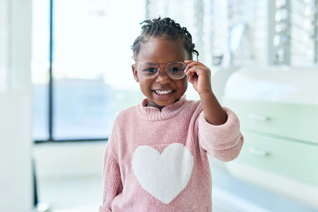 A young girl with braided hair wearing glasses and a pink sweater with a heart design smiles and playfully adjusts her glasses in a bright, modern setting.