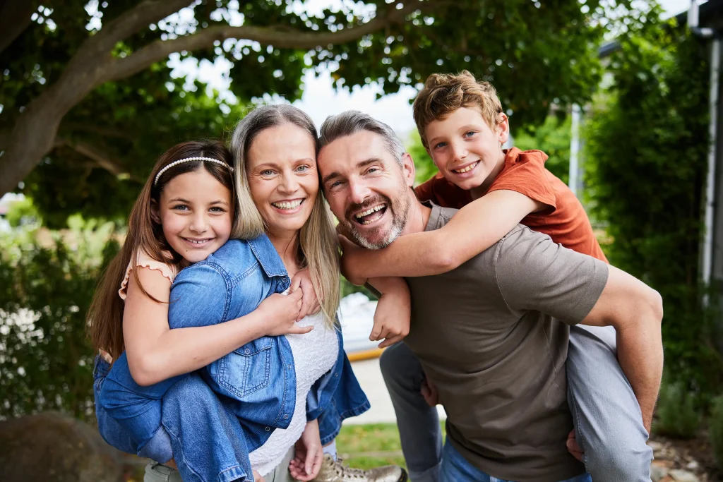 A happy family of four poses outdoors, with a mother and father each giving piggyback rides to a daughter and son, surrounded by greenery.