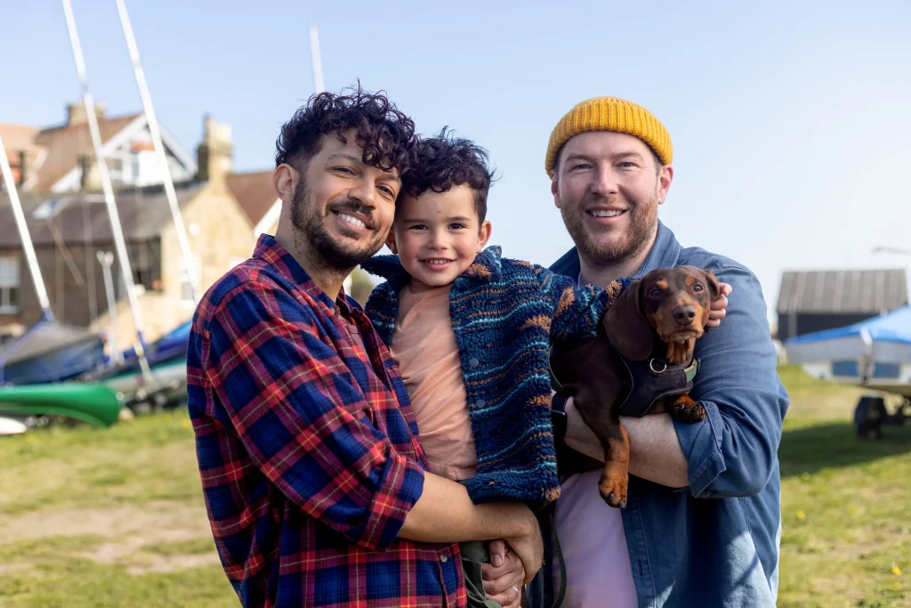 Two adult men and a child stand outdoors, smiling and holding a small dog. Sailboats and buildings are in the background.