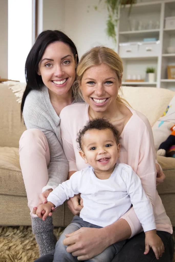 Two women hold a smiling toddler with curly hair in a neutral toned living room.