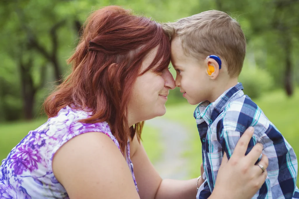 A woman with red hair smiles and presses her nose to the nose of a smiling child wearing a hearing aid. Green trees and grass are in the background.