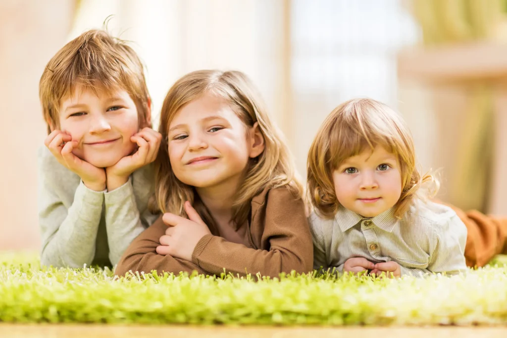 Three children lie on a green fluffy rug smiling at the camera