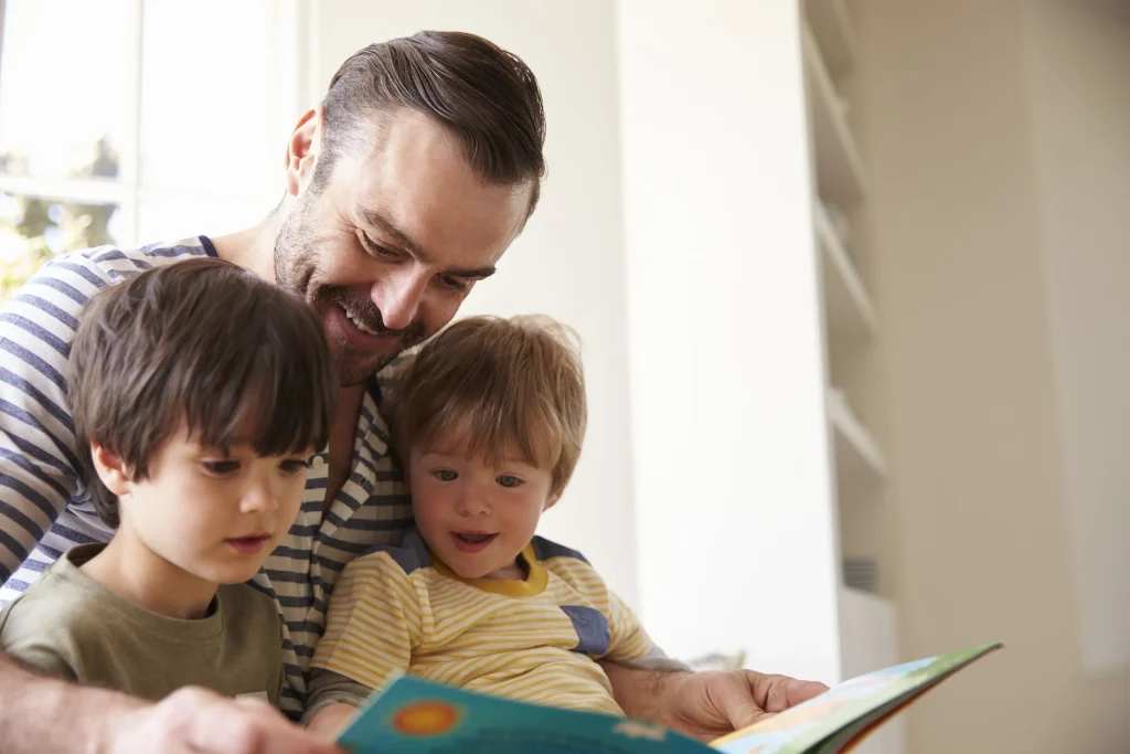 A man and two children are sitting in a window. The man is smiling and reading a book to the children.