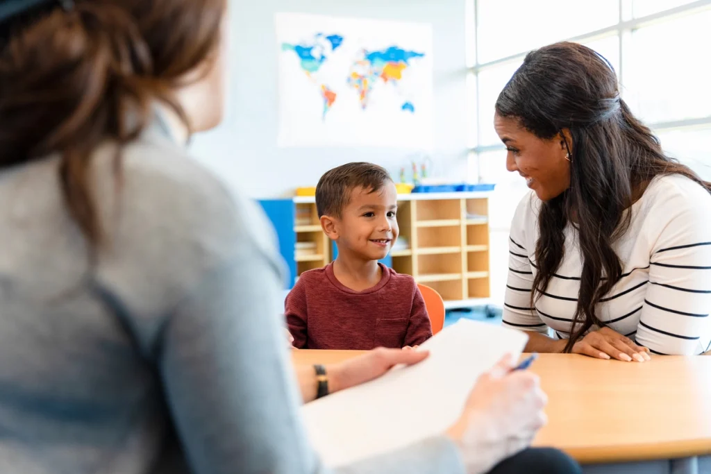 Two adult women sitting at a table in a classroom environment with a smiling child. One of the adults is writing on some paper.