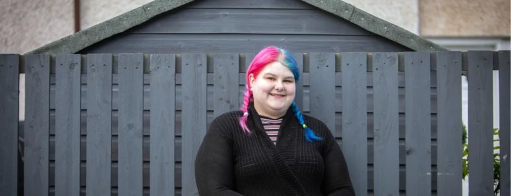 girl with blue and pink hair standing in front of a garden fence smiling to the camera